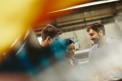 People in a tech environment discussing. Two males and one female. Caucasian (white) men and Asian woman. Primary color yellow. Secondary color blue.